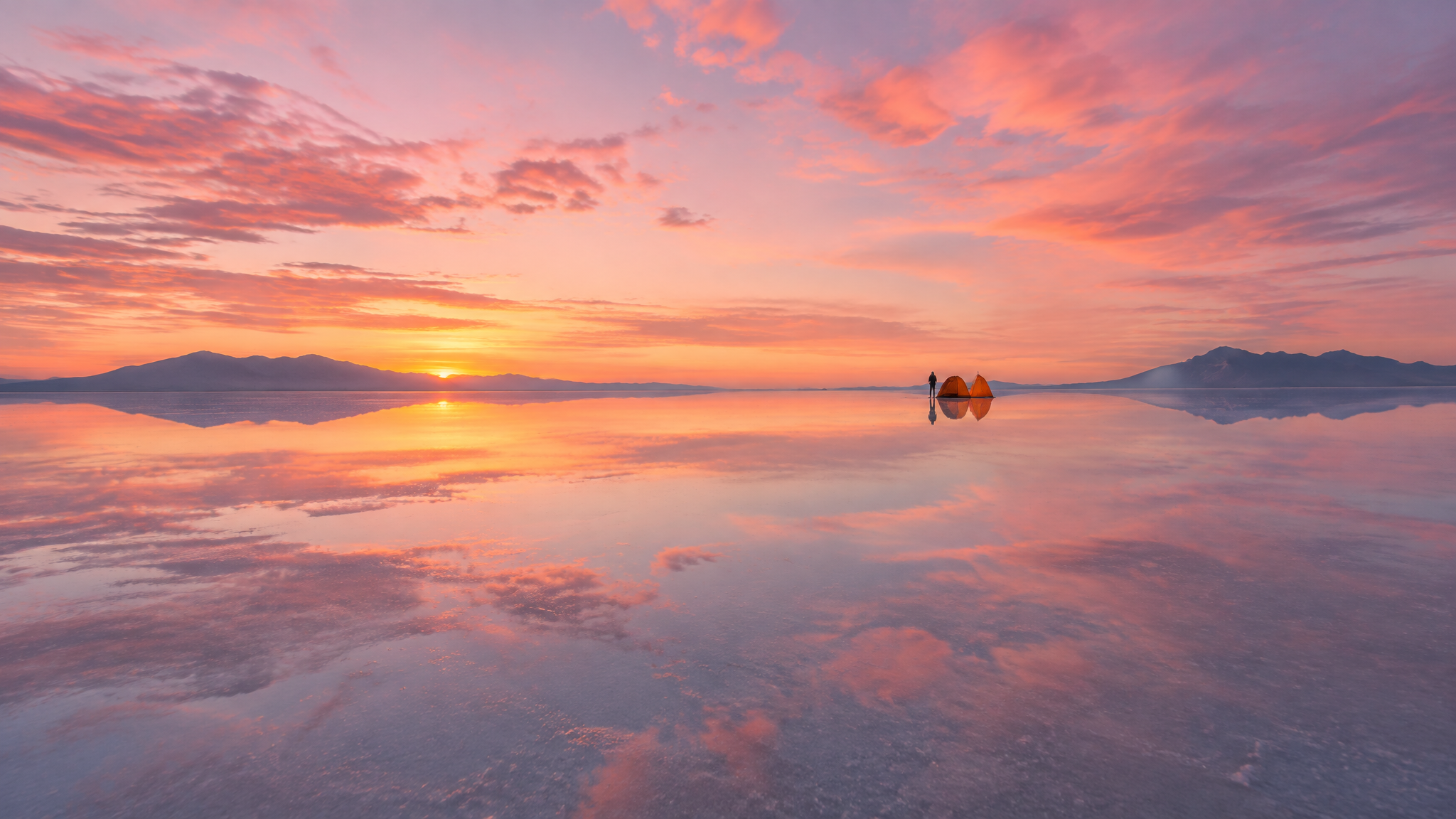 A single orange tent on a mirror-like salt flat reflecting a pink sunrise sky
