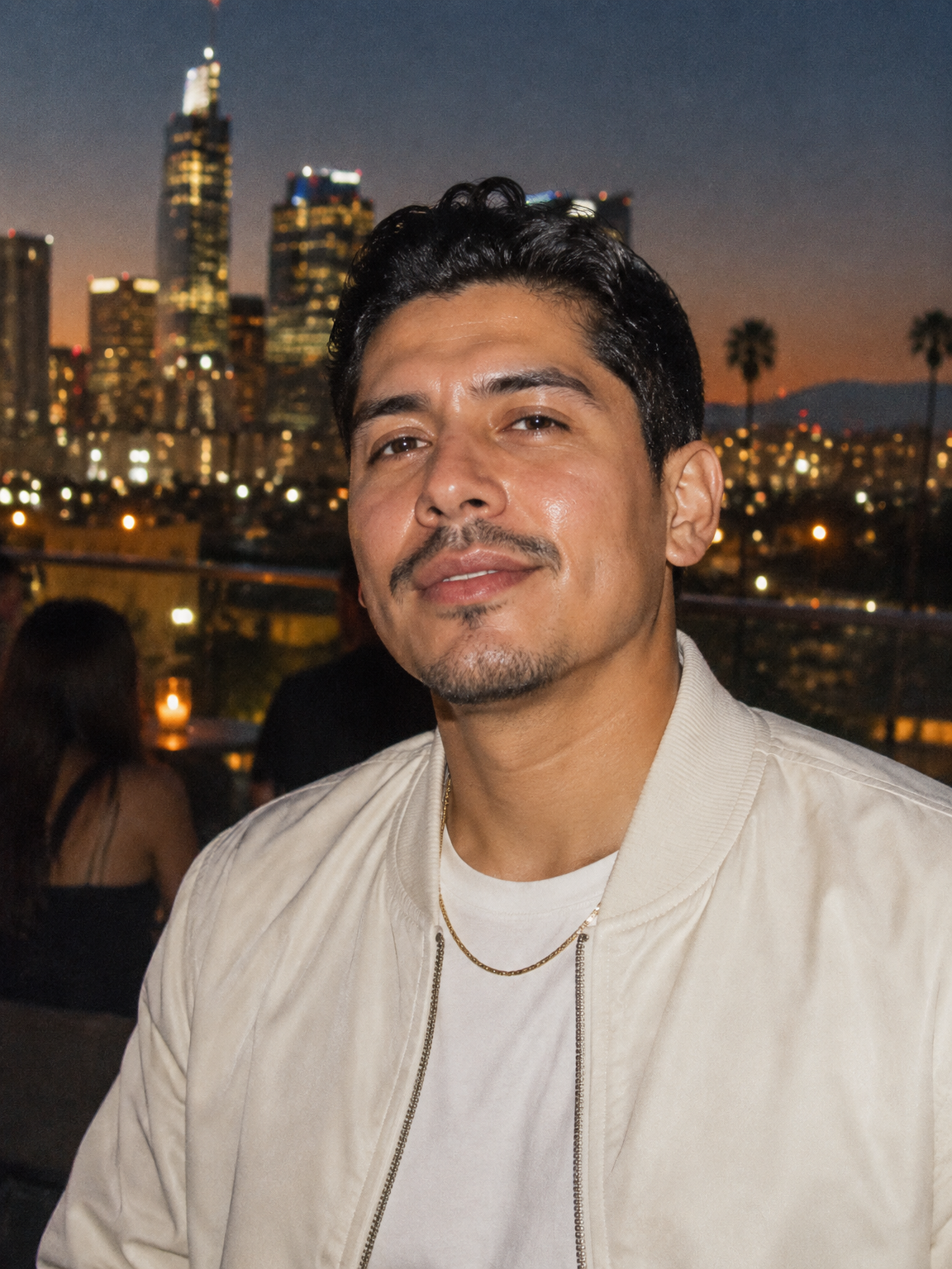 Latino man in a cream bomber jacket lit by direct flash on a rooftop bar with the Los Angeles skyline behind him