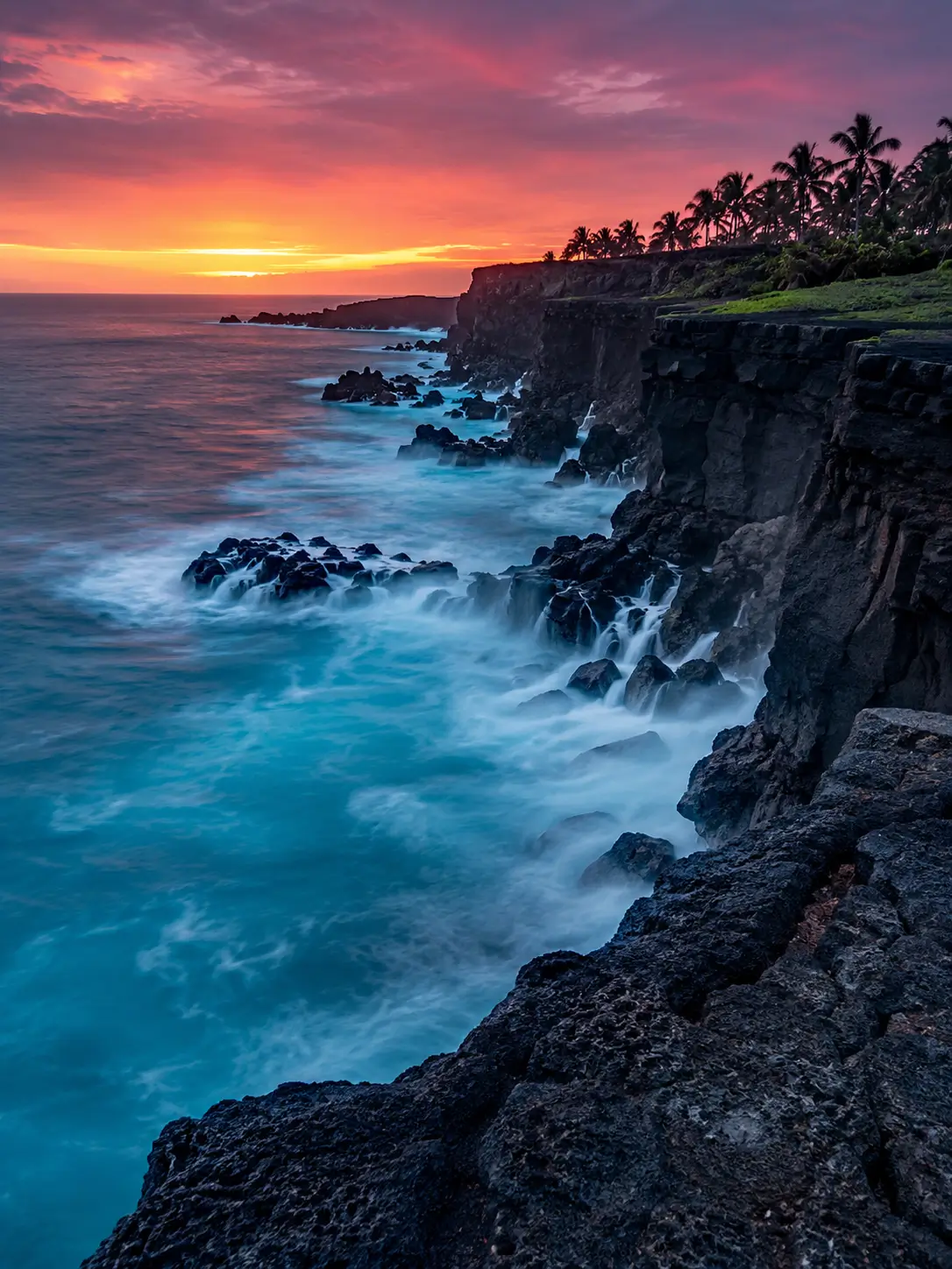 AI Arty Generator landscape of dramatic black volcanic cliffs, turquoise ocean waves and palm trees beneath a vivid orange and purple sunset.
