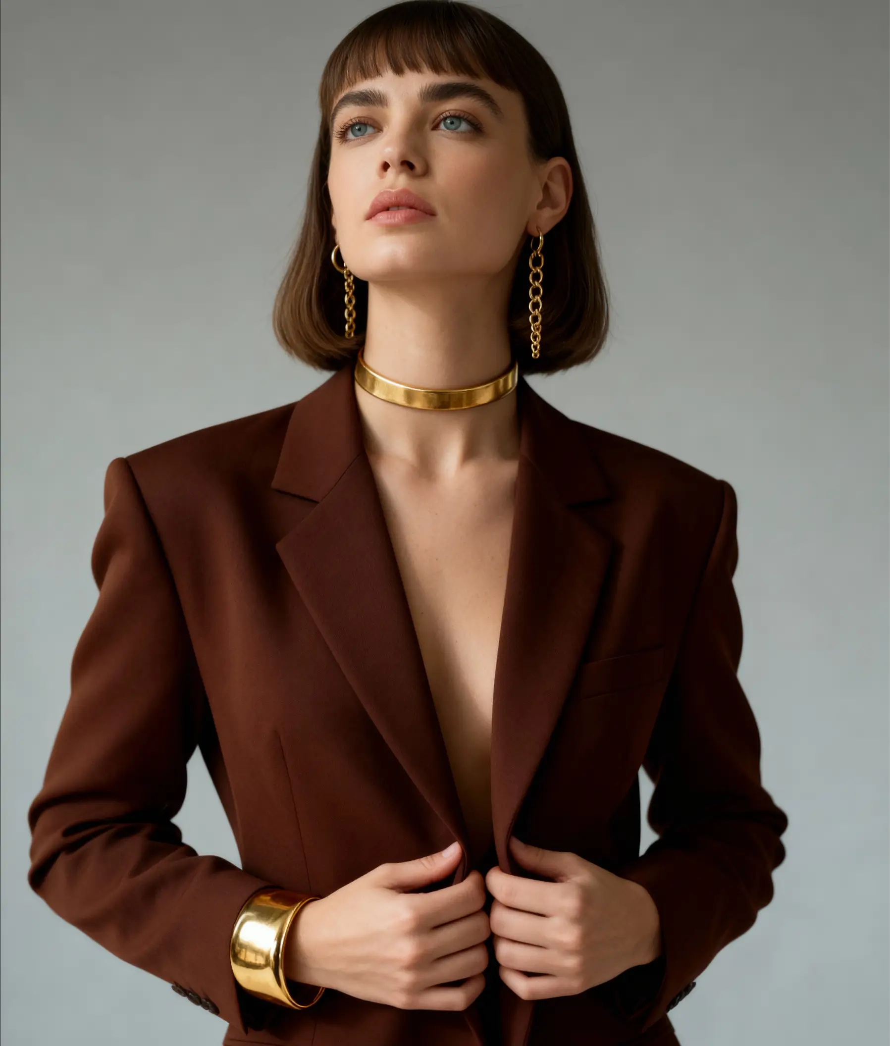 A professional headshot of a woman wearing a brown blazer with gold jewelry, posing elegantly against a soft gray background.
